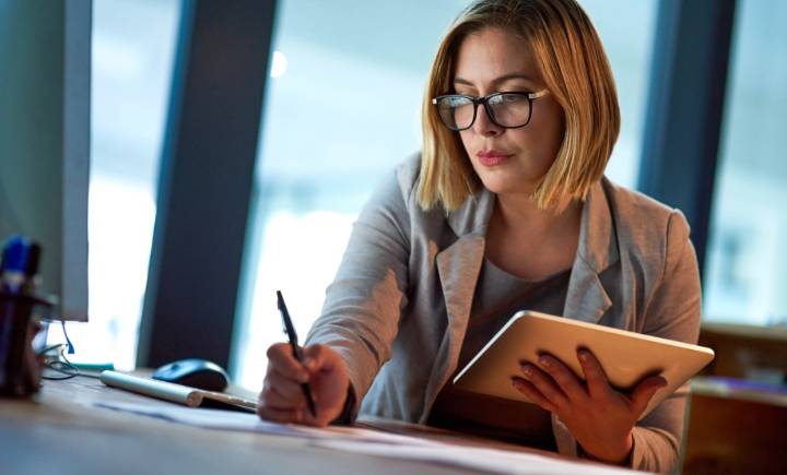 A woman wearing glasses works at a desk, holding a tablet in one hand and writing on paper with the other.
