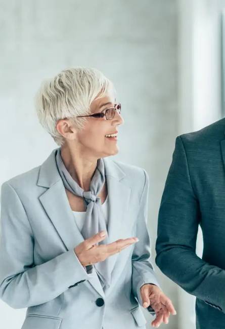 Older woman with short white hair and glasses, wearing a light gray suit and scarf, gestures while talking to someone beside her.