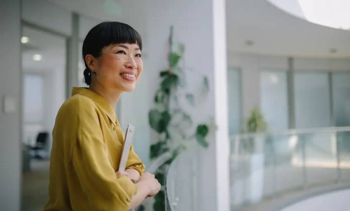 A woman in a yellow blouse stands indoors, holding a tablet and smiling, with a modern office background and a potted plant visible.