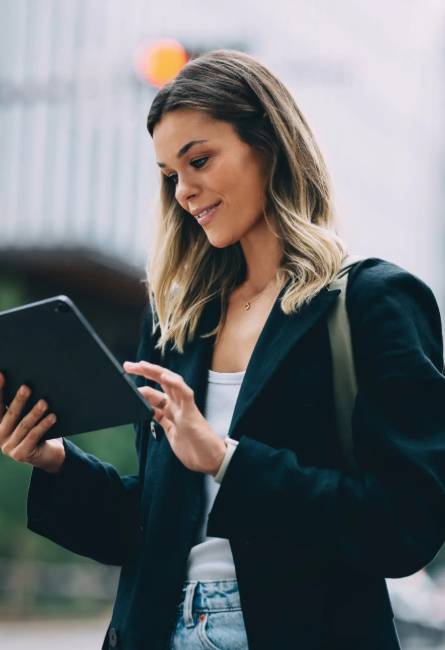 Woman wearing a black blazer and white shirt stands outdoors, smiling while using a tablet.