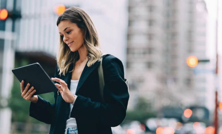 A woman standing outdoors in a city uses a tablet device while smiling, with blurred buildings and traffic lights in the background.