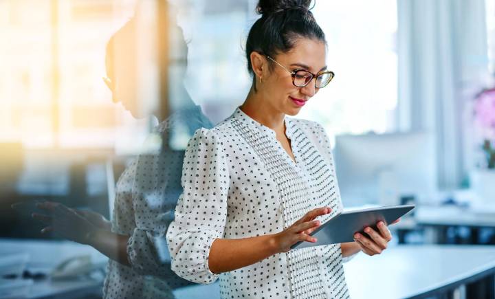 A woman in glasses and a white polka dot blouse uses a tablet in a bright, modern office with large windows.