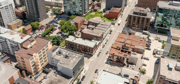 Aerial view of an urban street lined with multi-story buildings, parked cars, and small patches of greenery—an ideal backdrop for financial services industry events on a sunny day.
