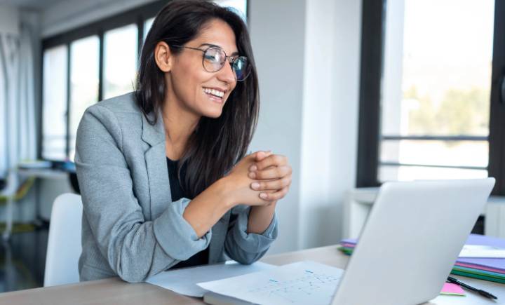 A woman wearing glasses and a blazer sits at a desk, smiling, and looks at a laptop in a bright office setting.