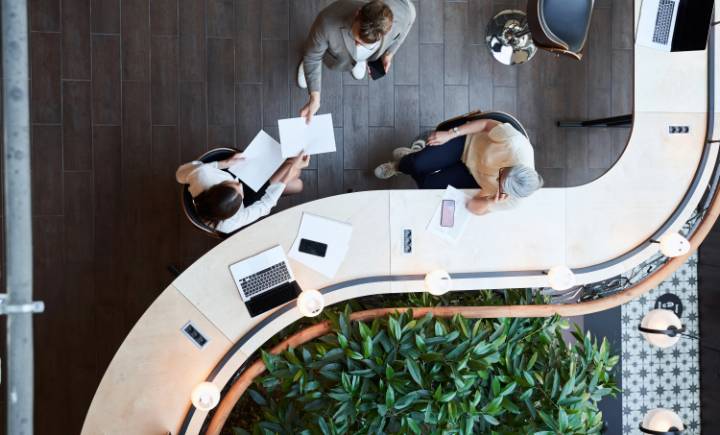 Three people sit and stand around a curved desk with laptops, papers, and plants visible, seen from above in a modern office setting.