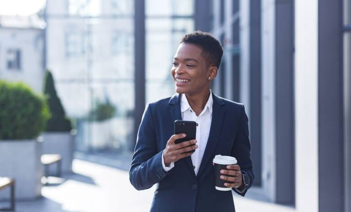 A person in a suit stands outdoors, holding a smartphone in one hand and a takeaway coffee cup in the other, smiling and looking to the side.