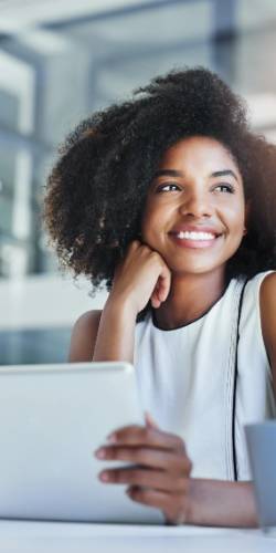 A woman with curly hair, dressed in white, smiles while seated at a desk and holding a tablet in a bright office setting.
