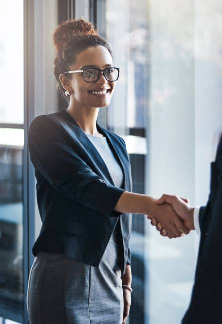 A woman wearing glasses and business attire smiles while shaking hands with another person in an office setting.