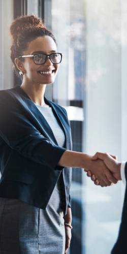 A woman in business attire, smiling and shaking hands with another person in an office setting, likely networking at financial services industry events.