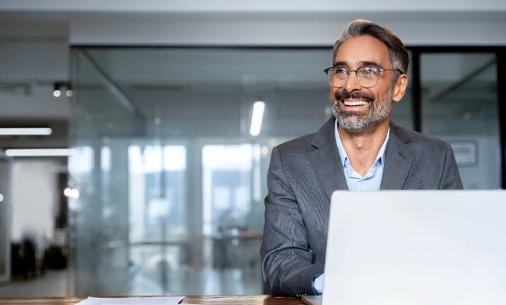 Middle-aged man with gray hair and beard wearing a suit, sitting at a desk with a laptop in a modern office, smiling and looking to his right.