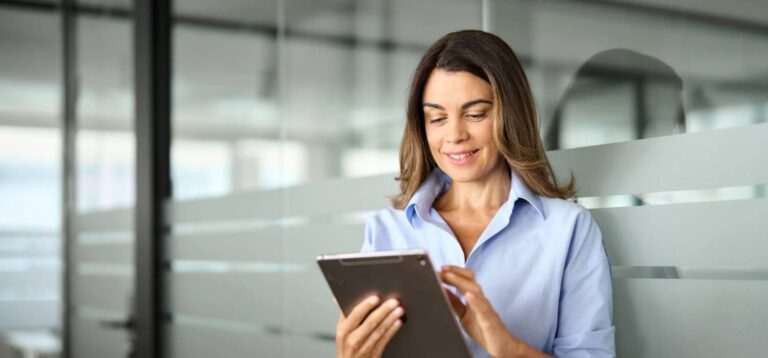 A woman in a light blue shirt stands indoors in front of glass panels, smiling while using a tablet.