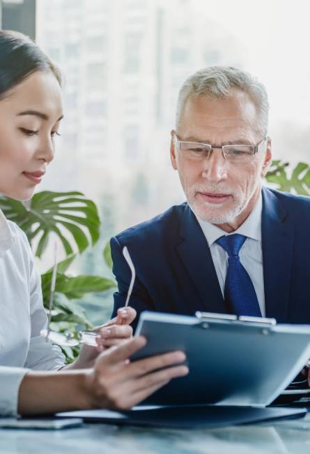Two business professionals, a woman and a man in formal attire, review documents together on a clipboard in a modern office setting.