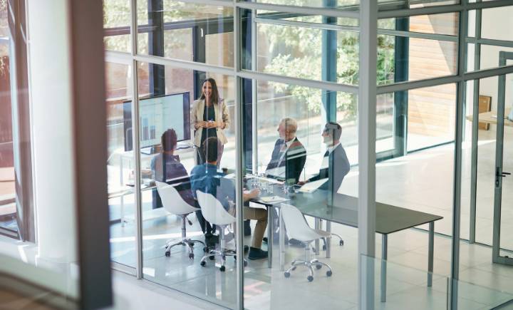 A group of business professionals sit around a conference table in a glass-walled office while a woman stands and presents at a screen.