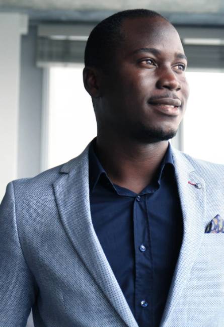A man in a light gray blazer and dark shirt stands indoors, looking out of a window with a neutral expression.