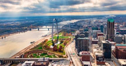 Aerial view of downtown St. Louis, Missouri, featuring the Gateway Arch, the Mississippi River, bridges, and surrounding cityscape under a cloudy sky.
