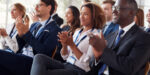 A group of professionally dressed people sit in rows, smiling and applauding, at what appears to be a business conference or seminar.