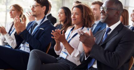 A group of professionally dressed people sit in rows, smiling and applauding, at what appears to be a business conference or seminar.