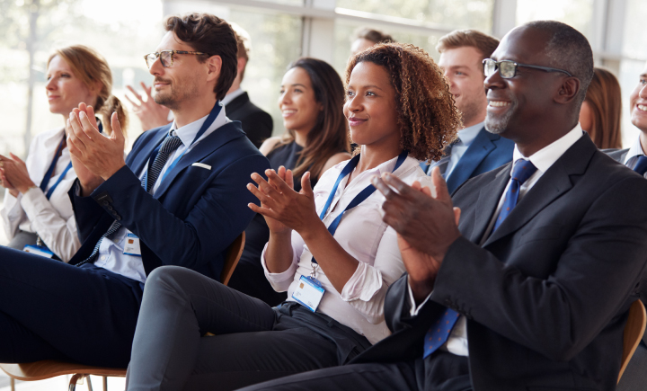 A group of professionally dressed people sit in rows, smiling and applauding, at what appears to be a business conference or seminar.