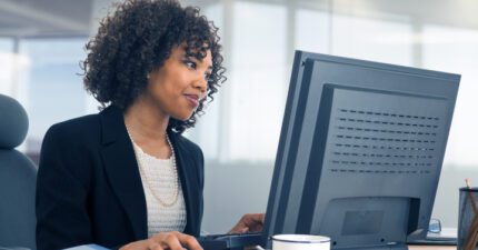 A woman in business attire works at a desktop computer in a modern office, with a coffee mug, notepad, and pen holder on the desk.
