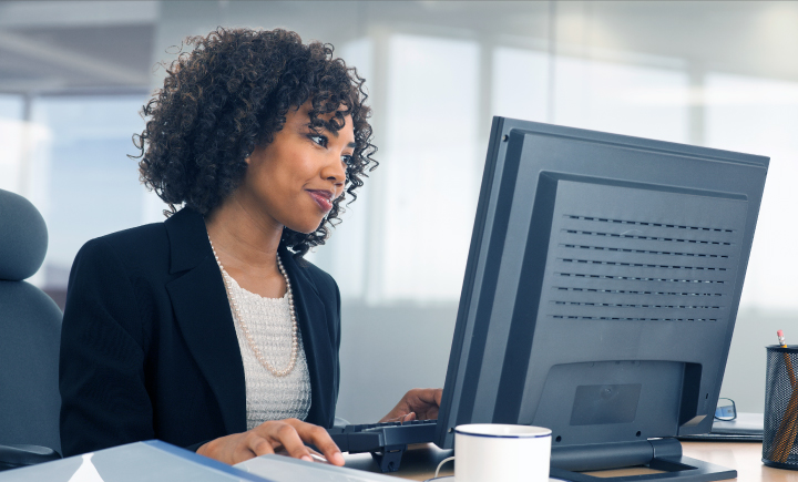 A woman in business attire works at a desktop computer in a modern office, with a coffee mug, notepad, and pen holder on the desk.