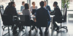 A group of people in business attire sit around a conference table having a meeting in a modern office with large windows.