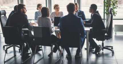 A group of people in business attire sit around a conference table having a meeting in a modern office with large windows.