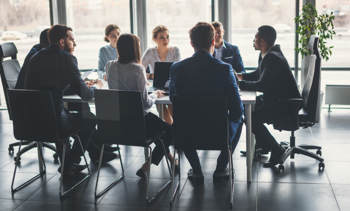 A group of people in business attire sit around a conference table having a meeting in a modern office with large windows.