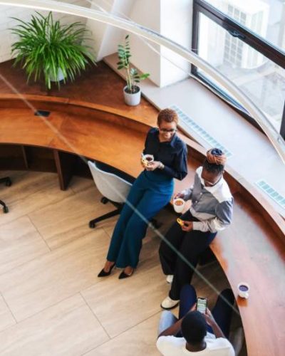 IStock 2152950751 Three people are gathered in a modern office lounge area, two standing and one seated, drinking beverages and conversing near a large window and potted plants.