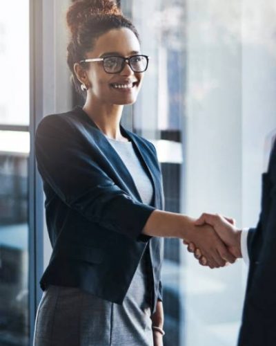 Shutterstock 2302260159 A woman wearing glasses and business attire smiles while shaking hands with another person in an office setting.
